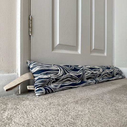 A draught excluder with a navy blue and white wavy pattern and a beige handle, resting on a carpeted floor in front of a white panelled door.