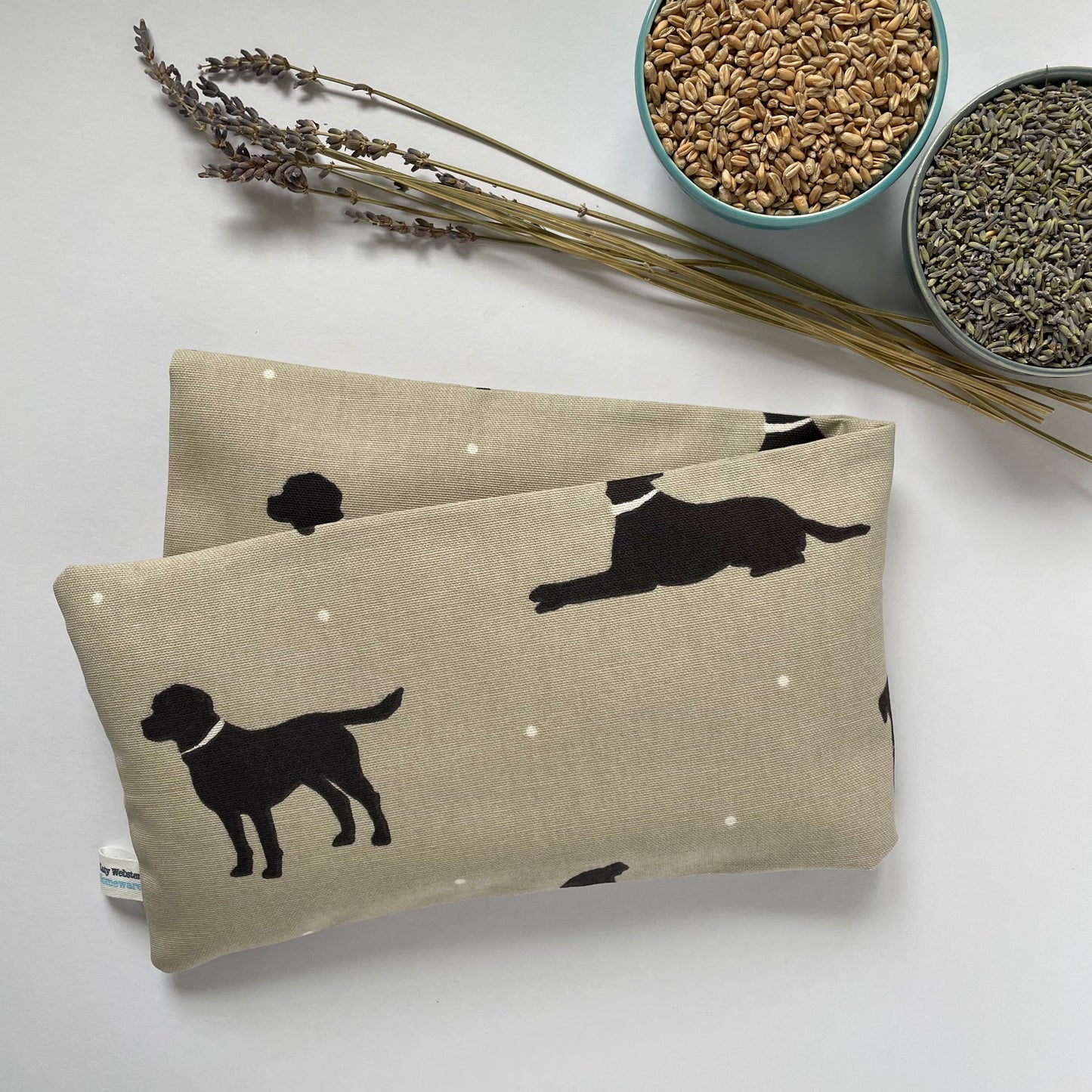 Two beige fabric pouches with black dog illustrations on a light surface, accompanied by dried lavender and bowls of seeds.