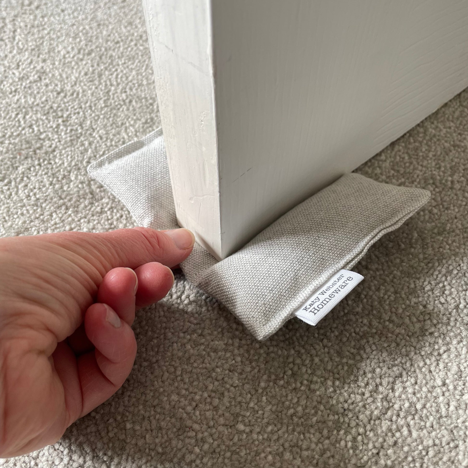 Fabric door stopper with a hand holding it against a white door on a carpeted floor.