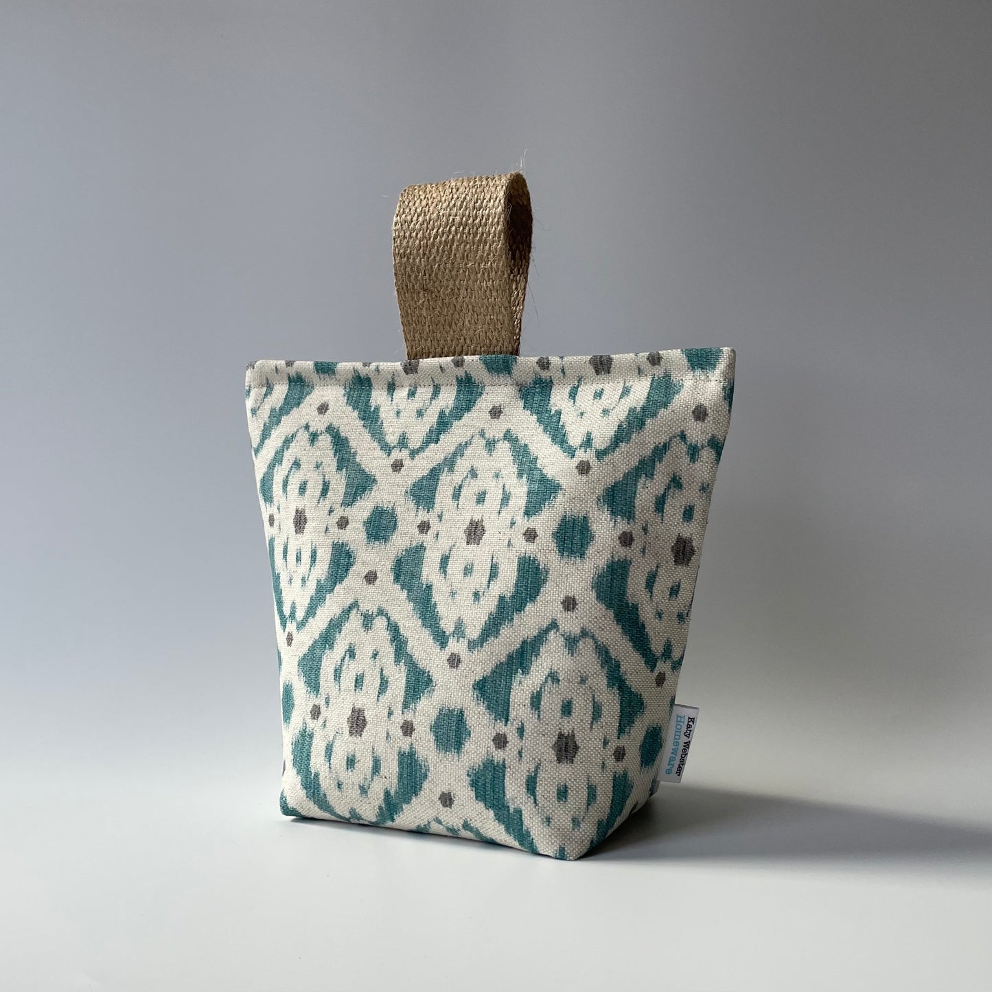 Fabric door stop with a neutral background and teal and grey geometric Ikat pattern with a thick jute handle on top.