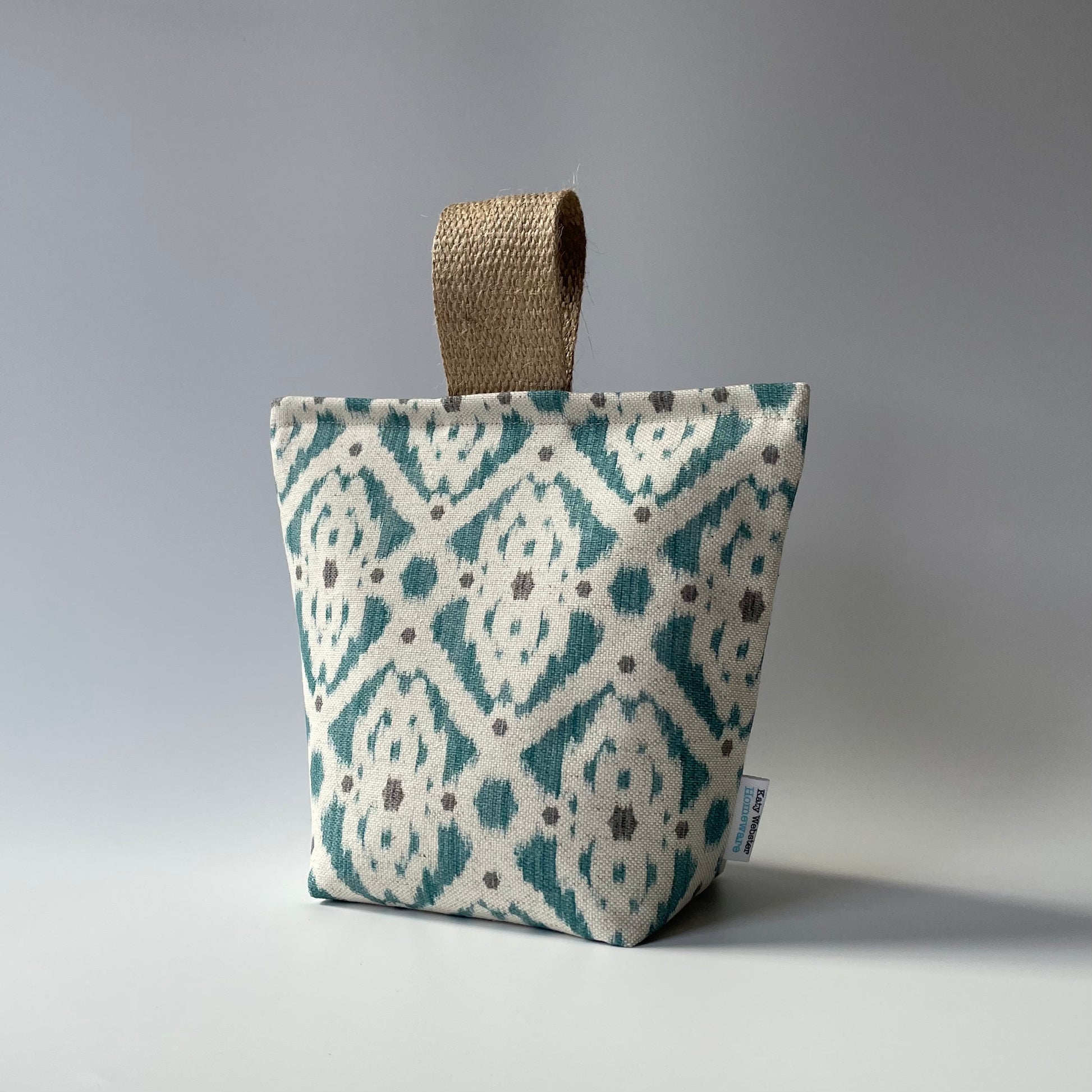 Fabric door stop with a neutral background and teal and grey geometric Ikat pattern with a thick jute handle on top.