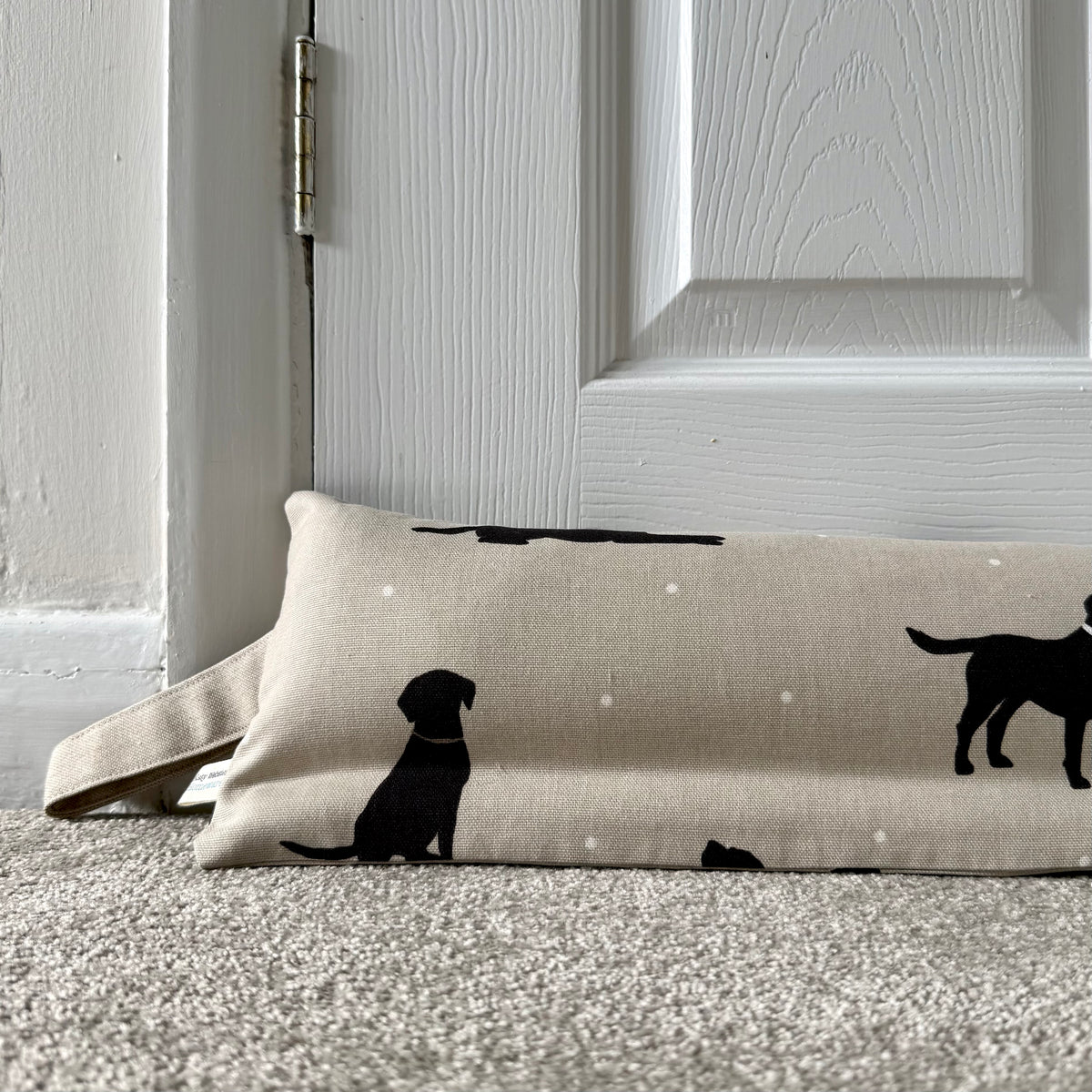 A beige draught excluder with black lab dog silhouettes and small white dots, placed on a carpeted floor in front of a white panelled door.