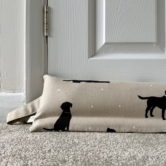 A beige draught excluder with black lab dog silhouettes and small white dots, placed on a carpeted floor in front of a white panelled door.