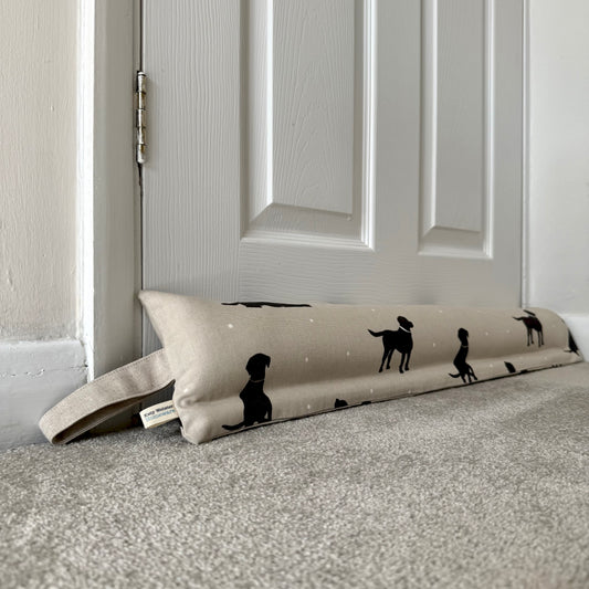 A draught excluder with a black lab dog silhouette pattern on a beige background, resting on a carpeted floor in front of a white panelled door.