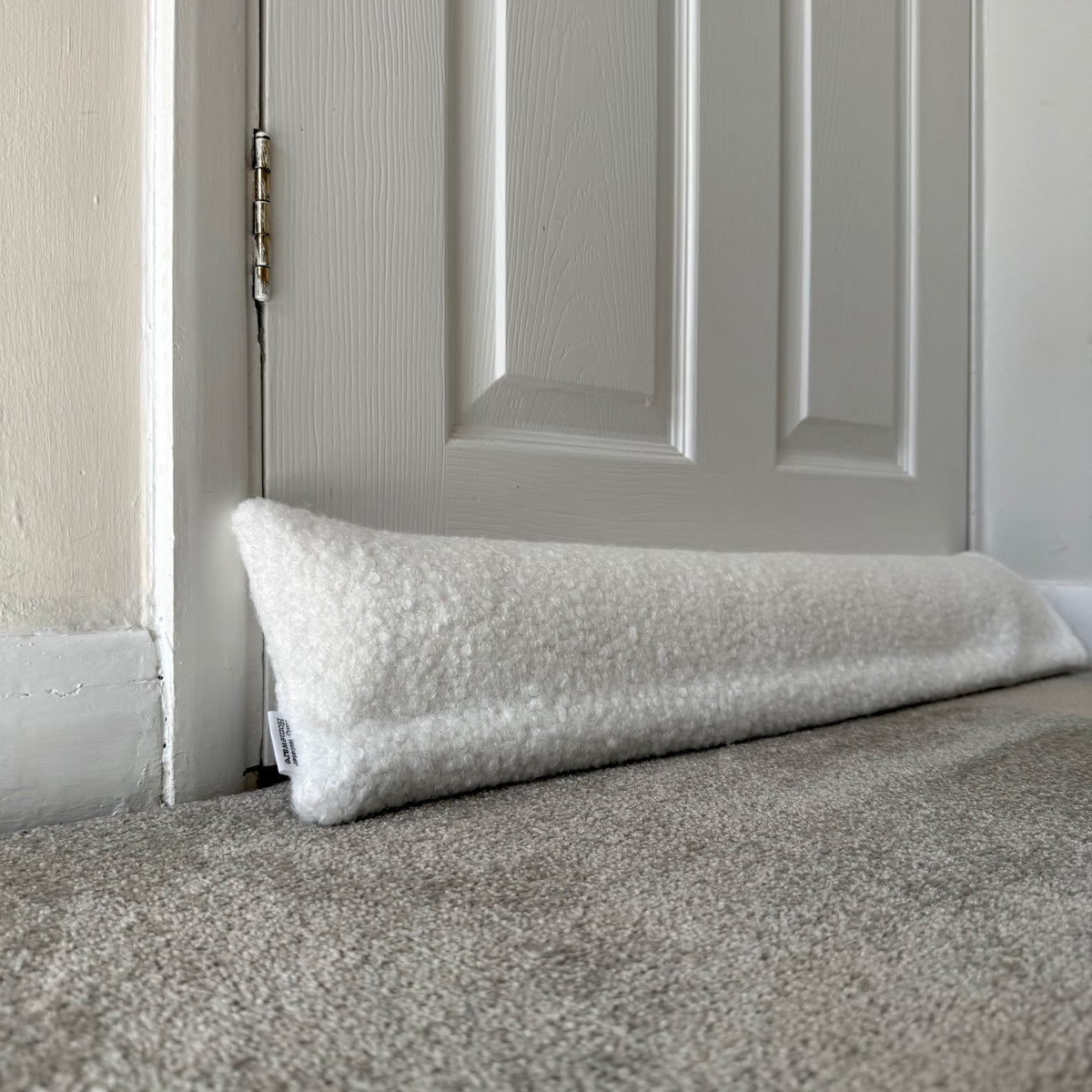 A white boucle draught excluder resting on a grey carpeted floor in front of a white paneled door.