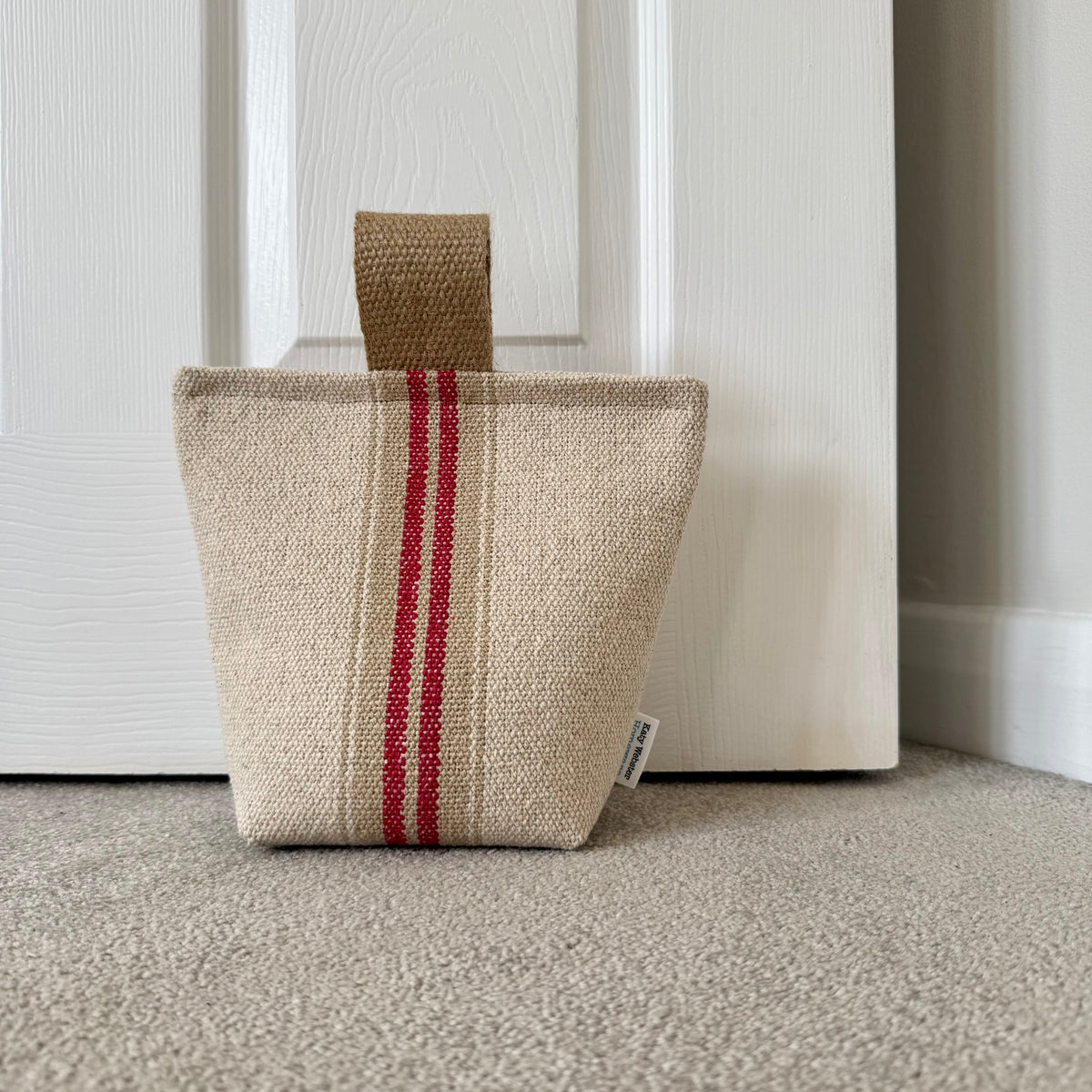 A woven grain sack fabric doorstop, featuring two vertical red stripes and a jute handle, standing on a carpeted floor in front of a white panelled door.