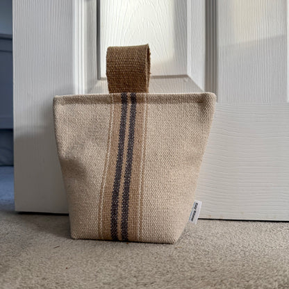 Close up of grey stripe grain sack fabric doorstop, featuring two vertical grey stripes and a jute handle, standing on a carpeted floor in front of a white panelled door.