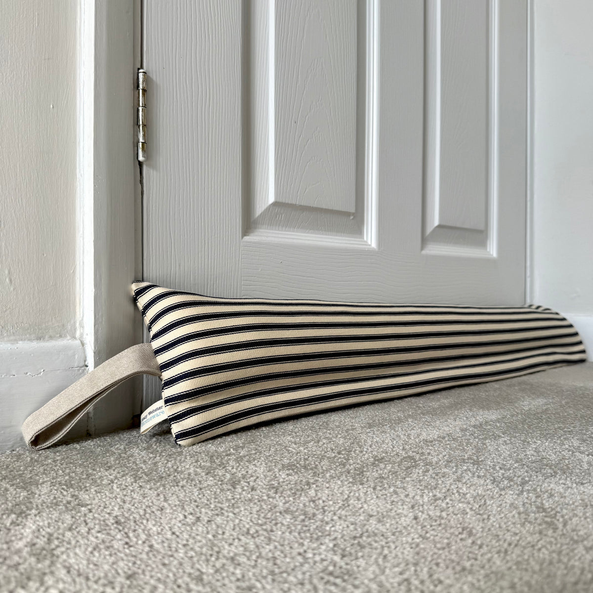 A striped navy and cream fabric draught excluder with a beige loop handle resting on a grey carpet in front of a white panelled door.