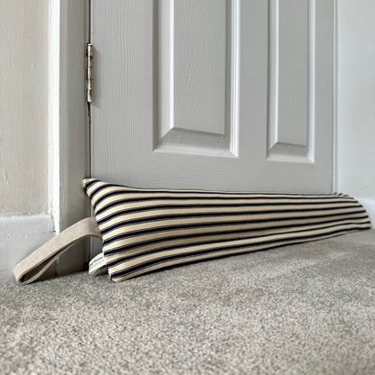 A striped navy and cream fabric draught excluder with a beige loop handle resting on a grey carpet in front of a white panelled door.