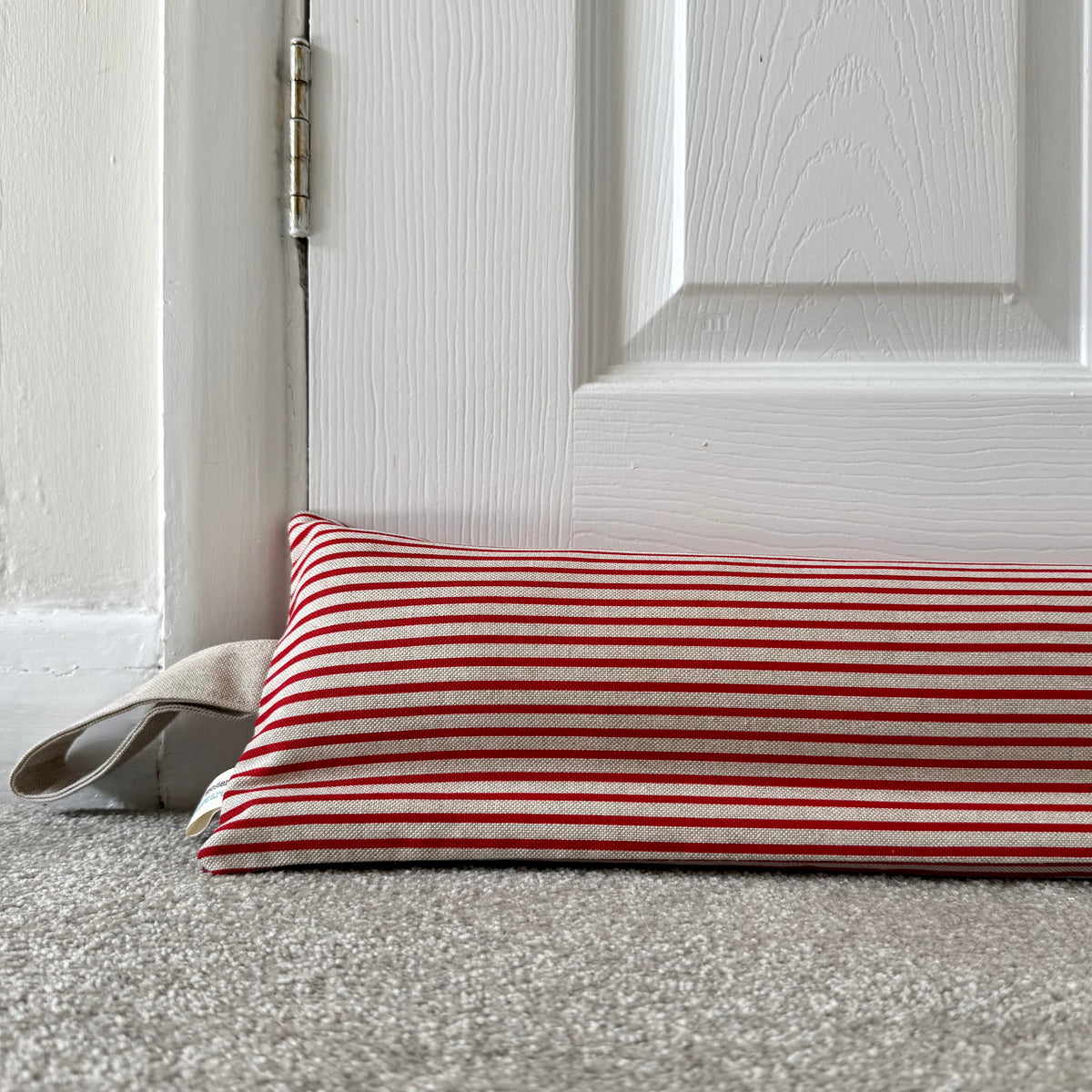 A weighted draught excluder with red and beige horizontal stripes, placed on a grey carpet in front of a white panelled door.