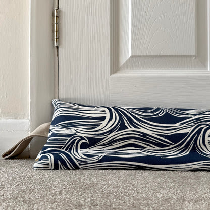 A weighted draught excluder with a navy blue and white wave pattern, placed on a carpeted floor in front of a white panelled door.