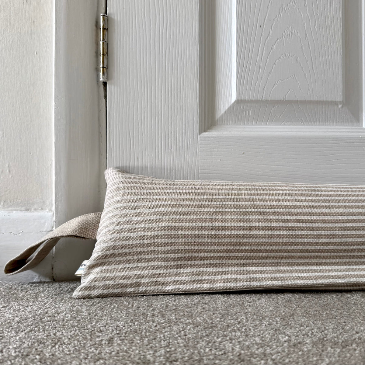A rectangular striped fabric draught excluder in beige and white lies on a gray carpet in front of a white paneled door.