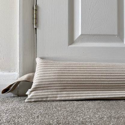 A rectangular striped fabric draught excluder in beige and white lies on a gray carpet in front of a white paneled door.